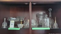 Full view of multiple cut crystal ware items arranged on wooden shelving, including two crystal bells, various bowls, decorative glasses, and containers.