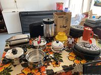 Array of kitchen items on floral tablecloth including Cuisinart Griddler, pots, roasters, carafe, and other containers.