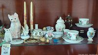 Wide shot showing the full lot of assorted decor items on a glass shelf including cat figurines, teacups and saucers, tall floral candles, and various ornamental containers