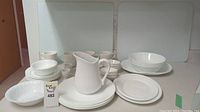 White porcelain dishware set including pitcher, plates, bowls, and mugs arranged on a table with two glass cutting boards in the background.
