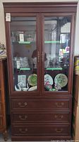 Full front view of tall dark wood display cabinet with glass doors showing items inside and three drawers below.