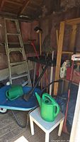 Photo of garden storage shed corner showing wooden ladder, garden tool organizer rack, multiple garden tools including rakes, two green plastic watering cans on a small white table, and a blue wheelbarrow.