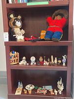 Wooden shelving unit with stuffed animals including teddy bear in red and blue, raccoon-like plush, Garfield toy, red and black mittens, and a variety of small home decor miniatures.
