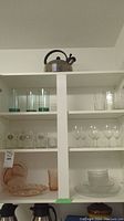 Wide view of shelf displaying glassware and glass dishes including tall glasses, wine glasses, and pink glass plates with bowls, plus a stainless steel stovetop kettle on top shelf.