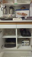 Kitchen cabinet open showing coffee urns, Pyrex measuring cups, various plates and kitchen tools in kitchen cabinet shelving