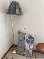 Photo showing a brass floor lamp with black lampshade next to the boxed WindChaser pedestal fan.