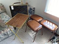 Photo showing three brown leather padded stools and one TV tray with brass folding frame and wood surface, plus carrying case with patterned exterior and leather handle.
