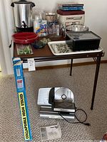 Full view of the lot arranged on and around brown card table showing meat slicer, cooler, kitchen tools, and other items.