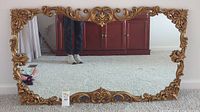 Full front view of large rectangular mirror on carpeted floor reflecting a wooden cabinet and part of the room.