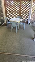 Full view of the round white resin patio table with four light blue resin chairs on concrete floor under a covered patio with wooden lattice wall.