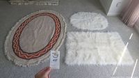 Three rugs displayed on carpeted floor: a large handwoven oval rug with fringe and multicolor zigzag border, a rectangular white faux fur rug, and a smaller white faux fur oval rug.