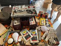 Overview of mixed lot of tools and home hardware items on a table, including saw set, gloves, drop sheets, electrical cords, bulbs, and hardware boxes.