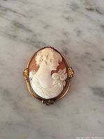 Front view of an oval cameo carved in shell depicting a woman profile with floral decoration, set in gold filled bezel with ornamental sides, photographed on marble background.