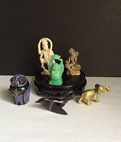 Six miniature figures including brass deity and elephant, celluloid blackbirds, lacquer trinket containers on wooden display stand with black background