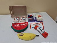 Photo of various games including two decks of Bicycle playing cards, Word-A-Melon pouch, banana-shaped Scrabble pouch, containers of red and white poker chips, and Scrabble Twist electronic game.