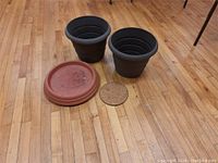 Two black plant pots, two terracotta-colored saucers, and a cork dolly on wooden floor