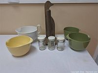 Three mixing bowls in yellow, white, and green, three glass storage jars with white lids, and wooden paper towel holder, all arranged on a white surface.