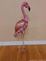 Full view of metal pink flamingo standing on wooden floor against beige wall, showing entire bird and base.