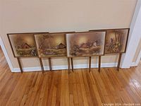 Set of four retro TV tables arranged side by side, showing framed countryside painted tops and four-legged wooden bases.