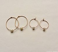Two pairs of gold hoop earrings each with a single cultured pearl displayed flat on a white background.