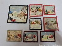 Overview of eight vintage hand made hooked rug coasters depicting winter and rural village scenes with houses, snow, and trees. Coasters are bordered in black or red fabric and vary slightly in size.