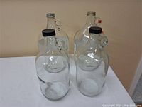 Four vintage clear glass vinegar jars with handles and varied caps, shown together on table against a wall; tallest is about 12 inches.