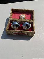 Two polished metal stress balls in an ornate fabric-covered decorative box with red lining and Chinese design.