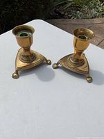 Pair of brass candle holders placed on white surface outdoors with visible sunlight and shadows. The holders have triangular bases with claw feet and show some surface patina.