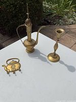 Photo showing all three brass items on white table outdoors: the genie bottle, candle holder, and decorative teapot piece.