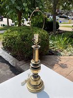 Brass lamp base on white surface near bush, daylight, showing full lamp with harp and electrical cord.