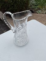 Crystal serving jug standing on a white surface, showing side view with handle and spout visible, highlighting the cut glass pattern.