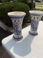 Pair of hexagonal Delft blue and white vases shown on white surface with greenery background.