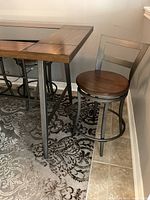 Photo showing bar height table corner with wood frame and glass center insert. One swivel stool with wood seat and metal legs positioned nearby.