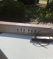 Photo of a modern ceiling light fixture with six evenly spaced light sockets and attached wiring, set on a table outdoors with visible garden in background.
