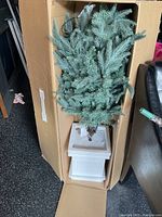 Photo of the artificial blue spruce tree inside its original box, showing the tree with green and blue-toned needles arranged in a white plastic pot base.