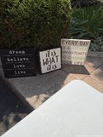 Three motivational wall art signs placed outdoors by bush and pavement, showing the full set.