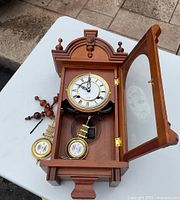Wooden pendulum clock with the door open, showing detached pendulum, bottom pegs, and clock face with Roman numerals.