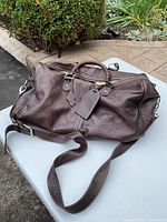 Full view of brown leather duffle bag with handles and shoulder strap laid on a white table outdoors.