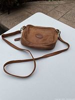 Brown leather Roots purse displayed on white surface showing strap and front embossed logo patch.