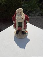 Photo of old-fashioned Santa Claus figurine standing on white surface outdoors, showing front and side angles with pipe in mouth.