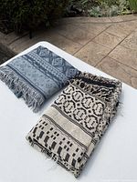 Two scarves folded on a white surface, one blue-grey with diamond and stripe patterns and fringe, one black-cream with geometric patterns and fringe
