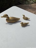 Three solid brass etched duck figurines displayed on a white surface outside, showing the different sizes and etching details.