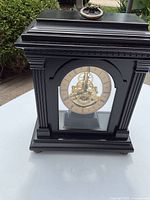 Front view of standing gear clock in black wooden case with visible brass gears and Roman numeral clock face.