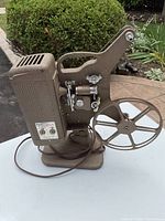 Side view of vintage Keystone Brightbeam K-70 film projector showing metal housing, reel holder, power cord, and adjustment knobs.