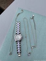 Silver-tone wristwatch with white and lilac link band next to two silver-tone necklaces, one with a lilac rectangular crystal pendant and one with a lilac crystal heart pendant.