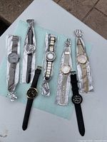 Seven assorted wrist watches laid out on a white surface, wrapped in protective plastic. Various styles including metal bands and strap designs.