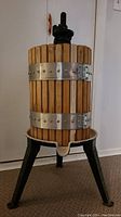 Front view of the antique wine press showing a wooden barrel with metal bands mounted on a cast iron tripod base with a screw press handle on top.