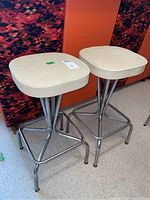 Pair of bar stools with cream vinyl padded seats and chrome metal bases, positioned on carpet with colorful background