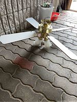 View of the ceiling fan on ground showing five white blades and four frosted glass light fixtures with brass motor housing.