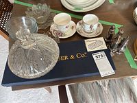 Crystal covered dish atop Godinger & Co. box, floral cups and saucers, salt and pepper shakers, and glass bowls on wooden table.
